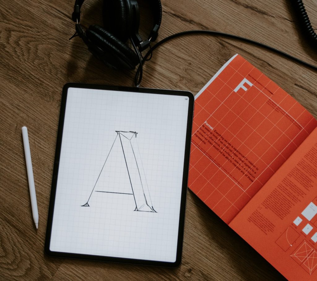 Image of a tablet with the letter A drawn on it. An apple pencil, headphones and a book are next to it.